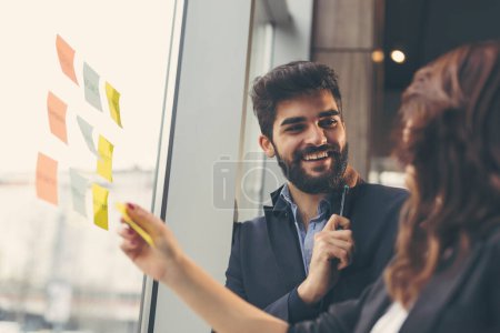 Téléchargez les photos : Homme d'affaires et femme d'affaires brainstorming utilisant des notes collantes sur un mur de verre dans un bureau - en image libre de droit