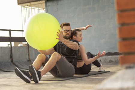 Téléchargez les photos : Couple assis dos à dos sur un tapis de yoga sur un toit-terrasse bâtiment, l'exercice en passant la balle pilates les uns aux autres - en image libre de droit
