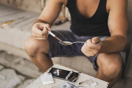 Foto de Un drogadicto intravenoso llenando una jeringa de heroína cociendo heroína de una cuchara, preparando su dosis de disparo - Imagen libre de derechos
