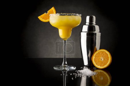 Foto de Cóctel de margarita de naranja con tequila, triple sec, zumo de naranja, hielo picado y un poco de sal en el borde de un vaso, decorado con una rebanada de naranja con coctelera al lado - Imagen libre de derechos