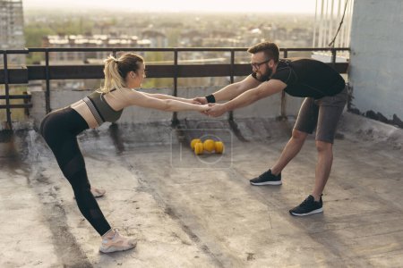 Photo for Young couple standing on a building rooftop terrace, holding hands and stretching before workout; urban skyline in the background - Royalty Free Image