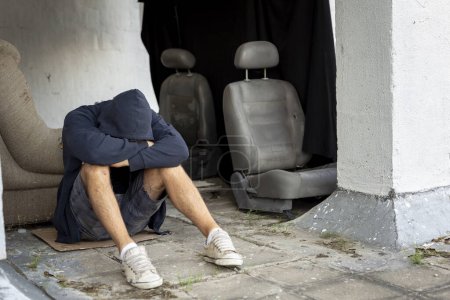 Foto de Deprimido, joven desesperado sentado en la terraza de la azotea del edificio, con capucha, sosteniendo la cabeza en las manos - Imagen libre de derechos