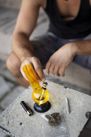 Foto de Hombre joven fumando marihuana usando pipa; detalle de la mano masculina llenando pipa con cannabis, preparándola para su uso. Enfoque selectivo en los dedos y la olla en la pipa - Imagen libre de derechos