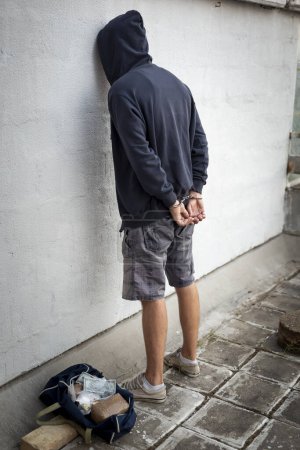 Téléchargez les photos : Un dealer en état d'arrestation, menotté et les mains sur le dos, debout à côté d'un mur. Focus sur la chaîne de menottes - en image libre de droit
