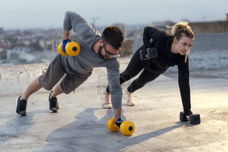 Photo for Couple working out on a building rooftop terrace, doing a plank exercise and lifting weights. Focus on the woman - Royalty Free Image