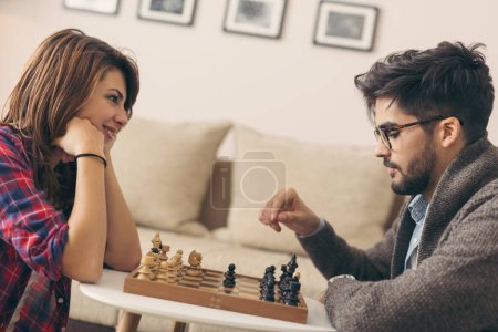 Téléchargez les photos : Couple amoureux assis sur le sol d'un salon, jouant aux échecs et s'amusant. Concentrez-vous sur le gars - en image libre de droit