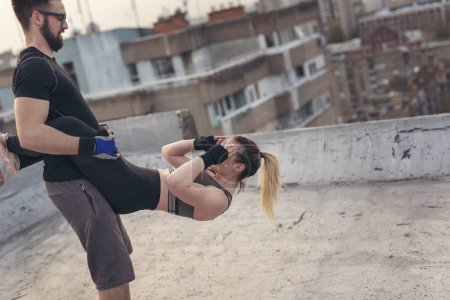 Foto de Pareja usando ropa deportiva haciendo ejercicios de crujidos colgantes en una terraza en la azotea de un edificio - Imagen libre de derechos