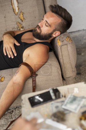 Téléchargez les photos : Drogué évanoui couché sur le canapé après avoir tiré avec de l'héroïne ; junkie surdosé avec injection intraveineuse d'héroïne - en image libre de droit