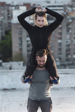 Foto de Pareja haciendo ejercicio en una terraza en la azotea del edificio, hombre cargando a una mujer sobre sus hombros y haciendo sentadillas - Imagen libre de derechos