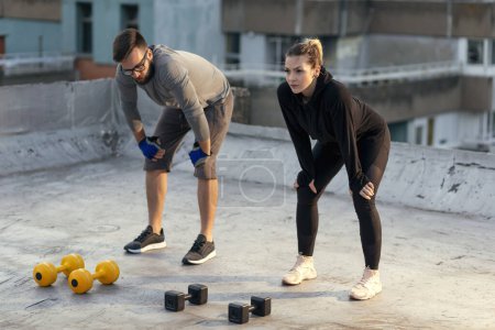 Photo for Couple taking a break from a building rooftop terrace workout, standing and holding hands on knees, stretching. Focus on the woman - Royalty Free Image