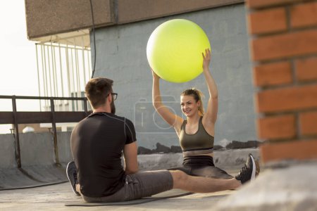 Téléchargez les photos : Couple assis sur un tapis de yoga sur un toit-terrasse du bâtiment, s'exerçant avec une balle de pilates - en image libre de droit