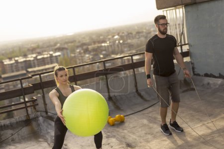 Photo for Couple working out on a building rooftop terrace, doing jumping rope and pilates ball squats. Focus on the girl - Royalty Free Image