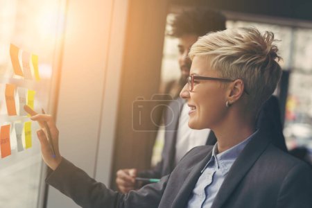Téléchargez les photos : Homme d'affaires et femme d'affaires brainstorming utilisant des notes collantes sur un mur de verre dans un bureau - en image libre de droit