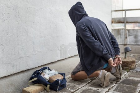 Foto de Narcotraficante bajo arresto confinado con esposas arrodilladas en el suelo. Enfoque en los bienes confiscados en la bolsa de lona - Imagen libre de derechos