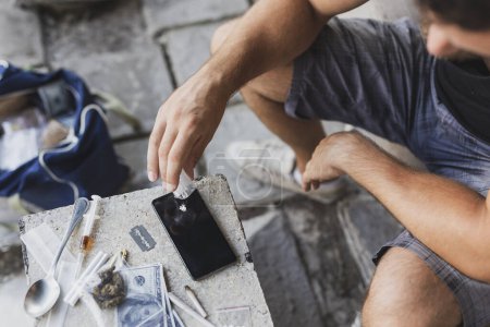Foto de Detalle de manos masculinas colocando heroína en una pantalla de teléfono inteligente; drogadicto oliendo heroína - Imagen libre de derechos