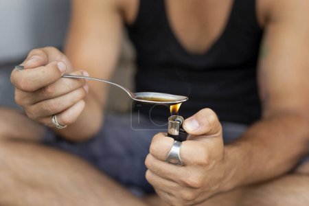 Foto de Detalle de manos masculinas cocinando heroína en una cuchara con un encendedor, preparándolo para uso intravenoso. Concéntrate en el encendedor y el pulgar - Imagen libre de derechos