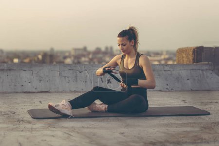 Téléchargez les photos : Belle jeune femme assise sur un tapis de yoga sur un toit-terrasse de bâtiment, enveloppant les mains dans des bandages, se préparant pour l'entraînement - en image libre de droit