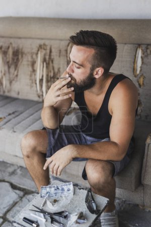 Téléchargez les photos : Jeune homme assis sur un canapé, relaxant et fumeur pot - en image libre de droit