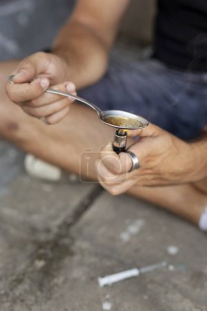Téléchargez les photos : Détail des mains masculines cuisinant de l'héroïne dans une cuillère avec un briquet, se préparant pour une utilisation intraveineuse. Concentrez-vous sur le bord de la cuillère et les bulles dedans - en image libre de droit