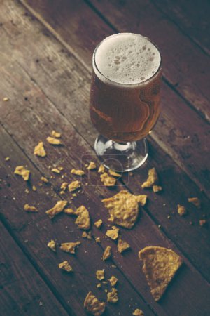 Photo for Glass of cold pale beer with some tortilla chips on a rustic wooden table. Selective focus on the foam - Royalty Free Image