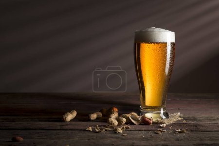 Foto de Un vaso de cerveza fría y ligera colocado en una montaña rusa de arpillera y unos cacahuetes sobre una mesa rústica de madera - Imagen libre de derechos