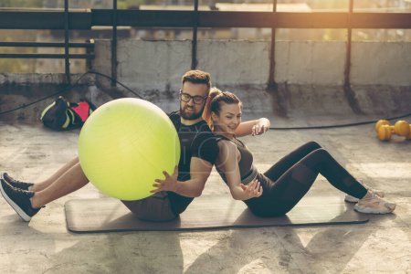 Téléchargez les photos : Couple assis sur un tapis de yoga sur un toit-terrasse du bâtiment, s'exerçant avec une balle de pilates - en image libre de droit
