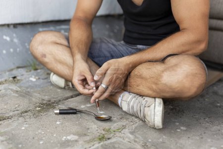 Téléchargez les photos : Un junkie intraveineux remplissant une seringue d'héroïne, se faisant cuire de l'héroïne à partir d'une cuillère, préparant sa dose de shoot up. Focus sur la seringue et les mains - en image libre de droit