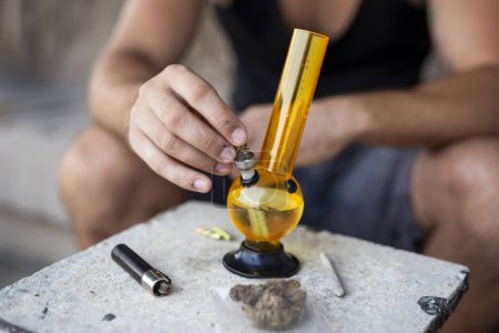 Photo for Young man smoking pot using bong; detail of male hand filling up bong with cannabis, getting it ready for use - Royalty Free Image