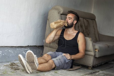 Téléchargez les photos : Ivrogne assis sur un toit-terrasse du bâtiment, tenant une bouteille dans un sac en papier, buvant - en image libre de droit