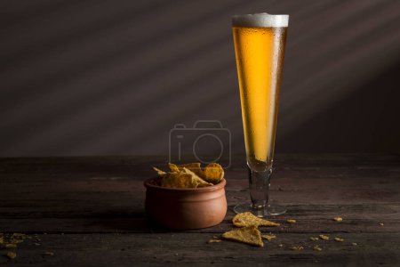 Foto de Vaso de cerveza fría pálida con un tazón de tortillas fritas sobre una mesa rústica de madera - Imagen libre de derechos