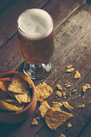 Téléchargez les photos : Verre de bière froide pâle avec un bol de copeaux de tortilla sur une table en bois rustique. Concentrez-vous sur la mousse - en image libre de droit