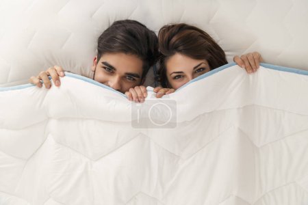 Téléchargez les photos : Vue du dessus d'un couple amoureux couché au lit, se cachant et regardant sous les draps - en image libre de droit