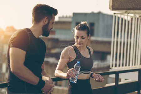 Photo for Young couple standing on a building rooftop terrace, drinking water and speaking while taking a break from a workout - Royalty Free Image