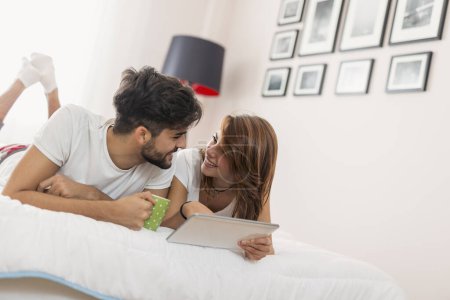 Photo for Couple in love, lying in bed, surfing the web on a tablet computer and having a cup of coffee - Royalty Free Image