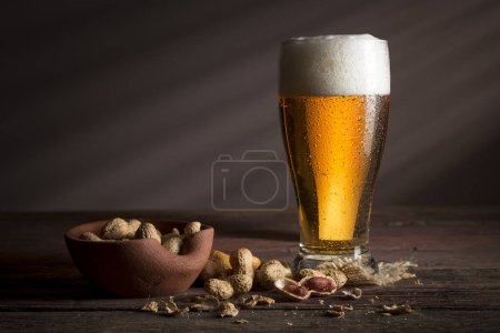 Photo for Glass of cold pale beer placed on a burlap coaster and a bowl of peanuts on a rustic wooden table. Focus on the froth - Royalty Free Image