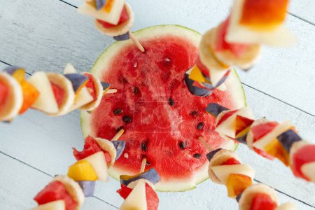 Téléchargez les photos : Plan de table d'une salade de fruits de saison mixte colorée servie sur des bâtonnets de barbecue, coincée dans une moitié de pastèque comme dessert de fête d'été - en image libre de droit
