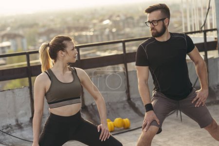 Photo for Young couple on a building rooftop terrace stretching before workout; urban skyline in the background. Focus on the girl - Royalty Free Image