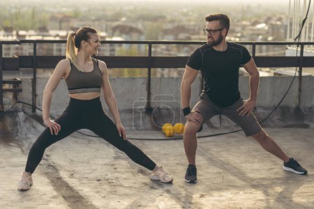 Photo for Young couple on a building rooftop terrace stretching before workout; urban skyline in the background - Royalty Free Image
