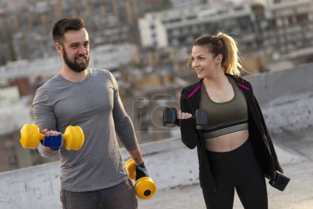 Photo for Young couple working out on a building rooftop terrace, lifting weights, doing arms exercises. Focus on the man - Royalty Free Image