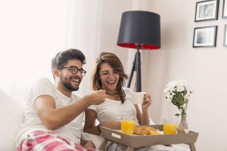 Photo for Happy couple in love lying in bed, having breakfast and enjoying their time together. Focus on the girl - Royalty Free Image