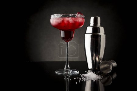 Téléchargez les photos : Cocktail de margarita aux cerises avec tequila, jus de cerise, glace concassée et un peu de sel sur le bord d'un verre, décoré de cerises. Cocktail shaker et jigger à côté - en image libre de droit