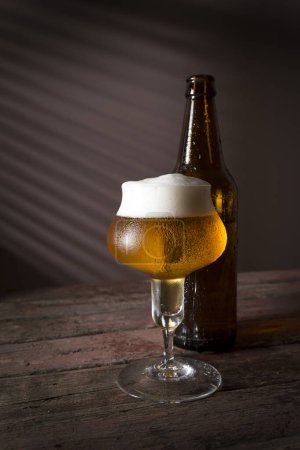 Téléchargez les photos : Verre de bière froide pâle avec une bouteille de bière en arrière-plan sur une table en bois rustique. Concentrez-vous sur la mousse - en image libre de droit
