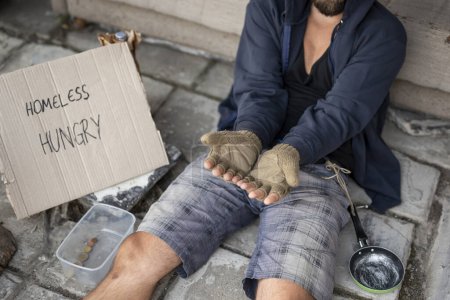 Photo for Beggar wearing hoodie over cap and fingerless gloves, sitting in the street, begging for money. Focus on the hands - Royalty Free Image