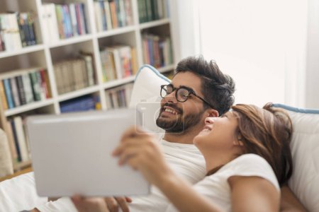 Téléchargez les photos : Couple amoureux, couché au lit, surfer sur le web sur une tablette, lire les infos - en image libre de droit