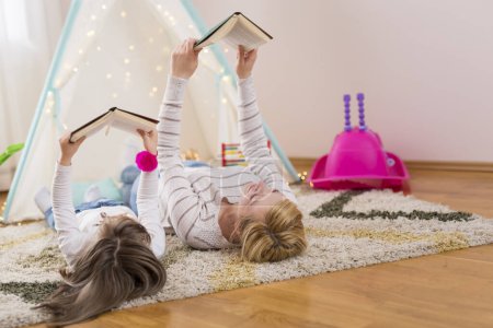 Téléchargez les photos : Mère et fille allongées sur le sol d'une salle de jeux, lisant des livres. Focus sur la mère - en image libre de droit