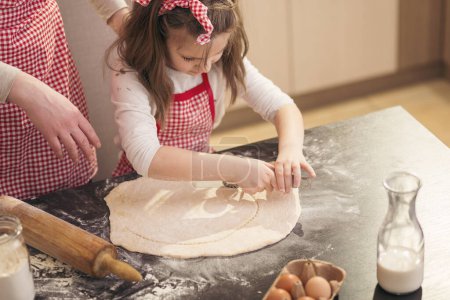 Foto de Hermosa niña con delantal en la cocina cortando masa para rollos con cortador de pizza con la ayuda de sus madres - Imagen libre de derechos