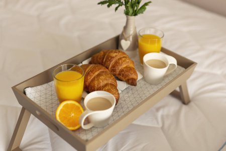 Téléchargez les photos : Vue en angle élevé d'un plateau de petit déjeuner avec vase de fleurs, jus d'orange, croissants et café placés sur le lit. Focus sélectif sur le croissant gauche - en image libre de droit