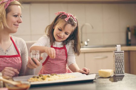 Photo for Mother and daughter enjoying making pizza in the kitchen; daughter adding oregano on top before baking. Focus on the daughter - Royalty Free Image