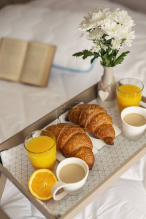 Téléchargez les photos : Vue en angle élevé d'un plateau de petit déjeuner avec vase de fleurs, jus d'orange, croissants et café placés sur le lit. Focus sur le croissant - en image libre de droit