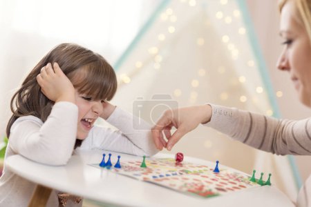 Foto de Madre e hija sentadas en una sala de juegos, jugando un juego de ludo; madre reposicionando el peón - Imagen libre de derechos